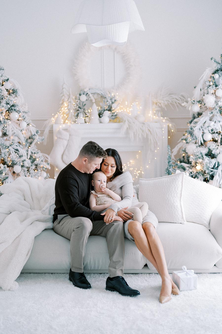 Family sitting on a white couch in a beautifully decorated living room with Christmas trees, wreaths, and glowing lights in the background.