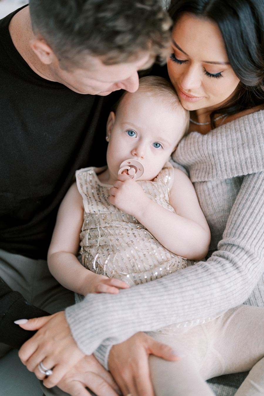 A loving family moment featuring a close-up of a toddler with a pacifier, dressed in a sparkly outfit, embraced by two adults in cozy sweaters, symbolizing warmth and affection.