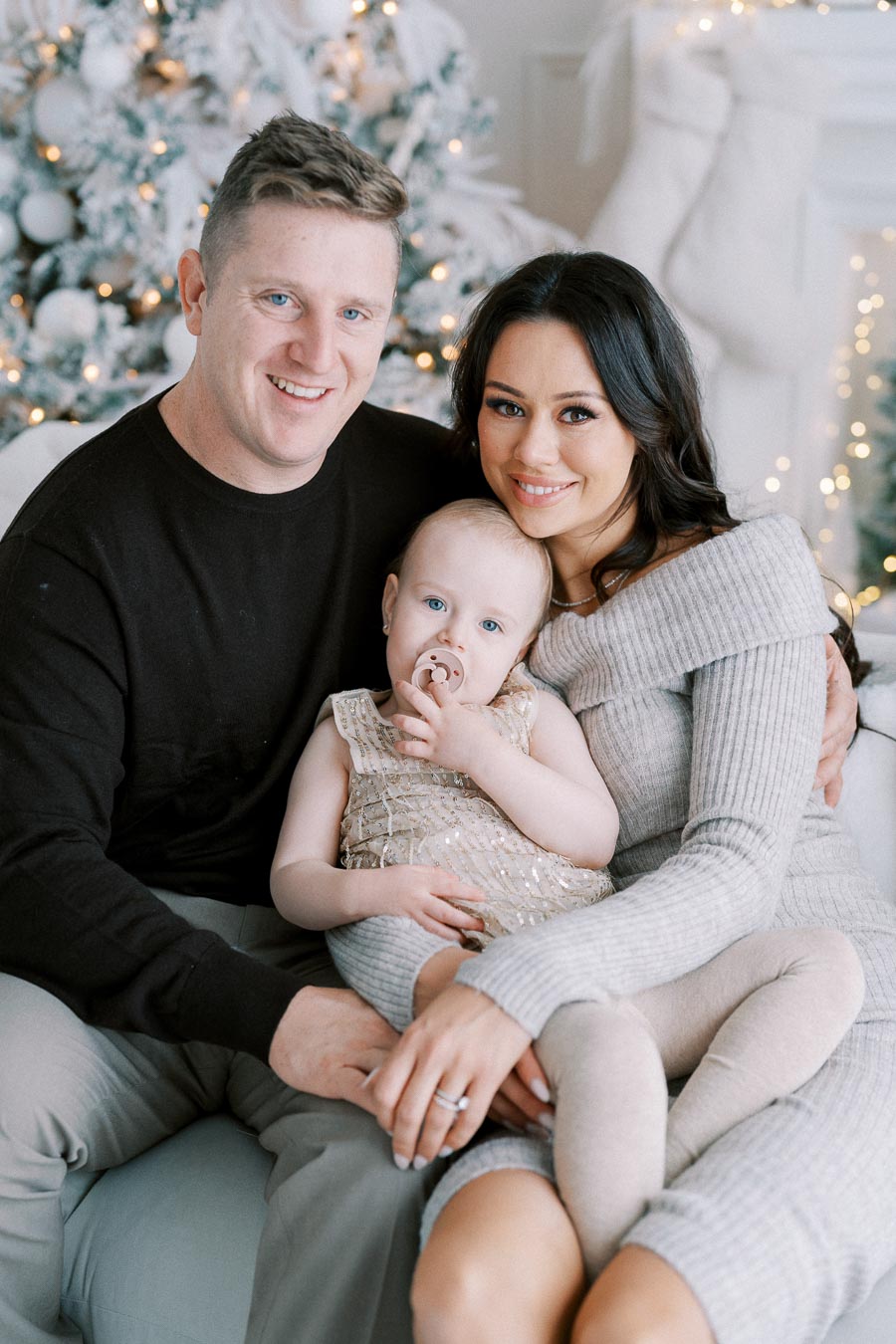 A happy family sitting together in front of a decorated Christmas tree, with the father in a black sweater, the mother in a gray off-shoulder dress, and the baby wearing a gold dress and holding a pacifier.