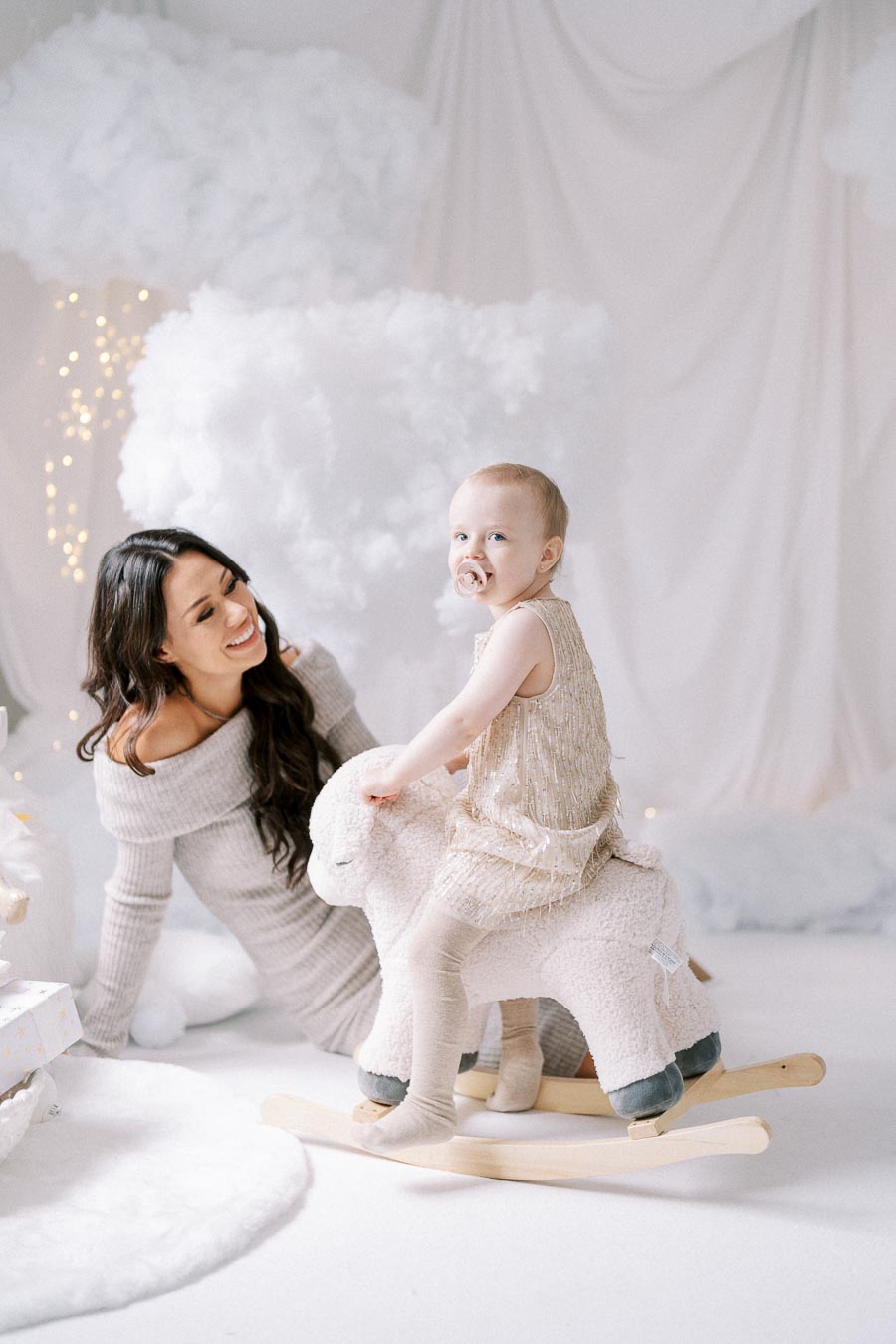 A smiling woman in a gray dress kneels beside a baby, dressed in a sparkly outfit, sitting on a plush rocking sheep in a bright, cloud-themed room decorated with fairy lights.