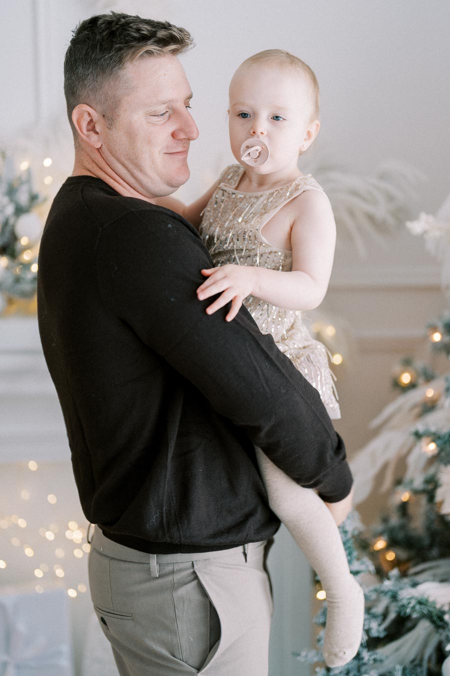 Father holding toddler with pacifier near decorated Christmas tree.