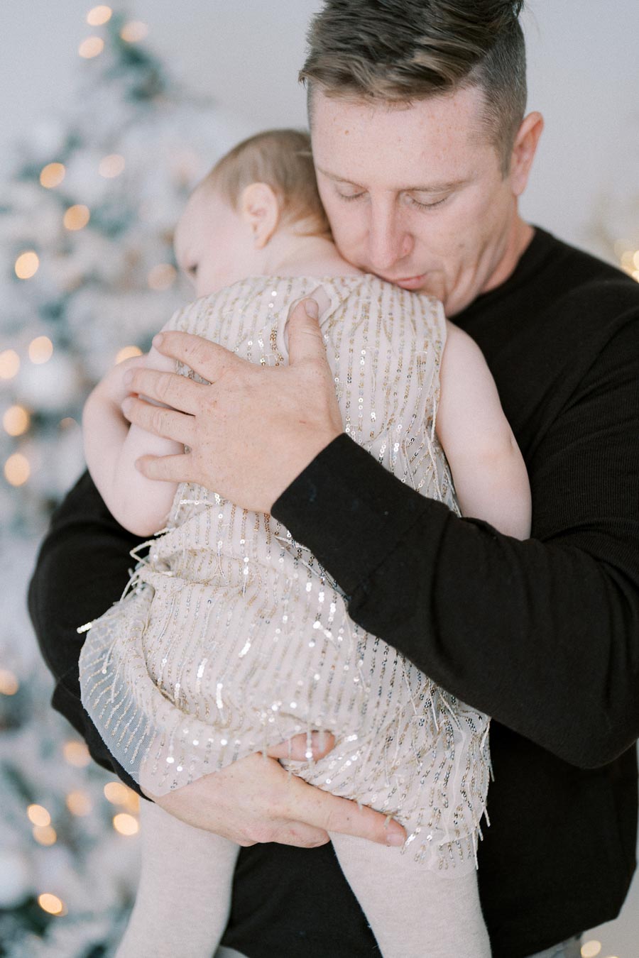 A man gently holding a baby in a sparkly dress with a decorated Christmas tree in the background, conveying warmth and holiday spirit.