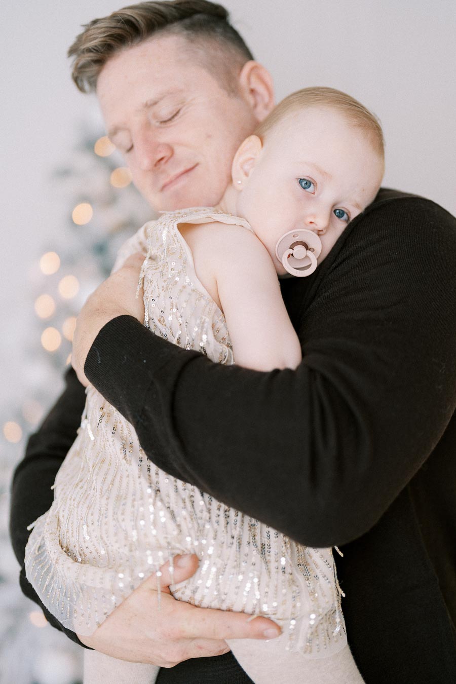 Alt text: Father holding baby with pacifier, dressed in a sparkly gown, in front of a softly lit background.