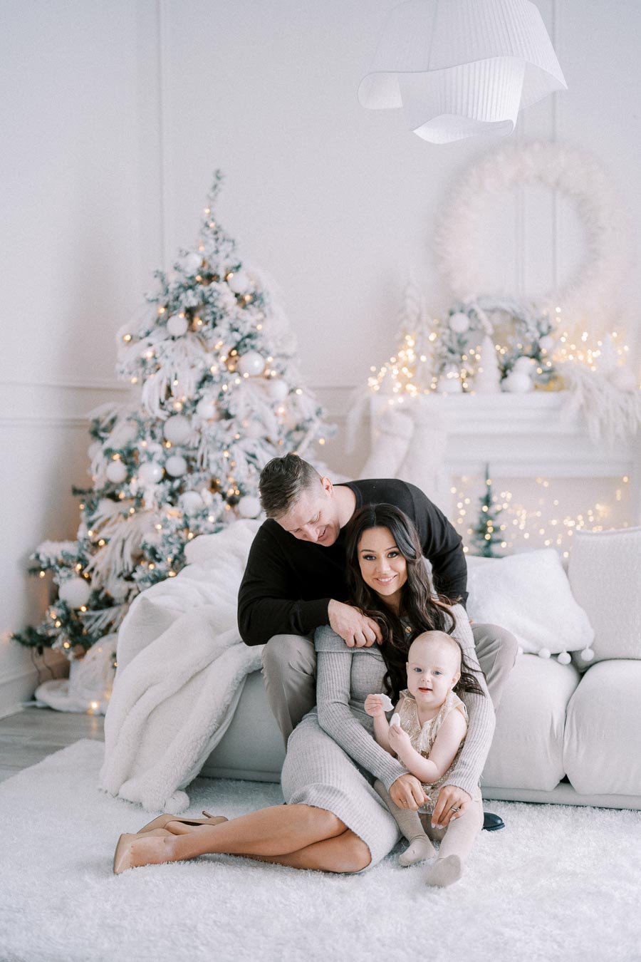A family sitting together in a cozy, white-themed living room decorated for Christmas, with a beautifully lit tree and festive decor in the background.