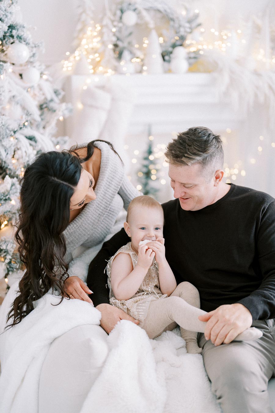 A happy family consisting of a mother, father, and baby sitting together on a cozy couch, surrounded by festive Christmas decorations and a brightly lit tree, showcasing a warm holiday atmosphere.