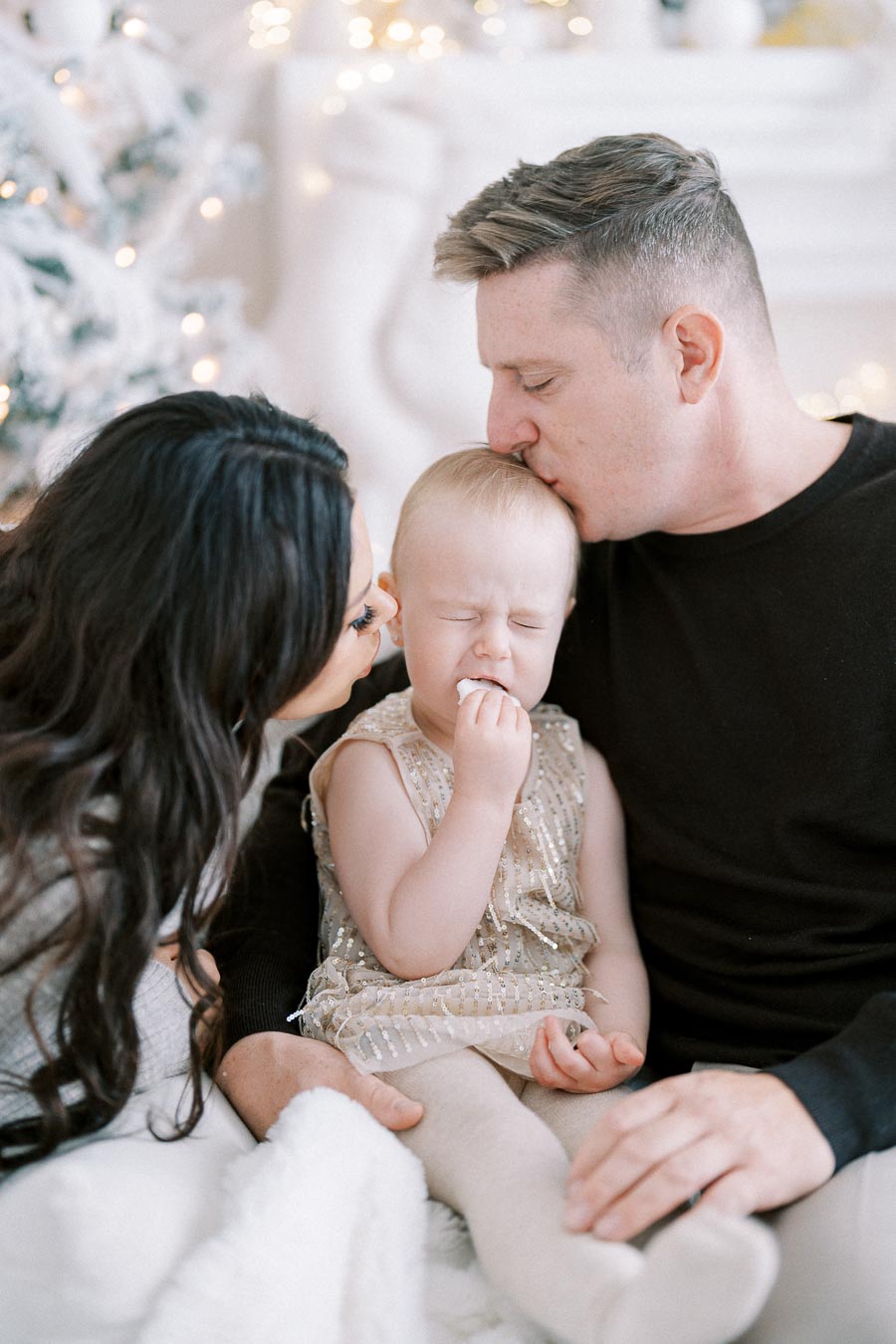 Family holiday moment with parents kissing their child, sitting on a cozy couch beside a decorated Christmas tree, conveying love and warmth.
