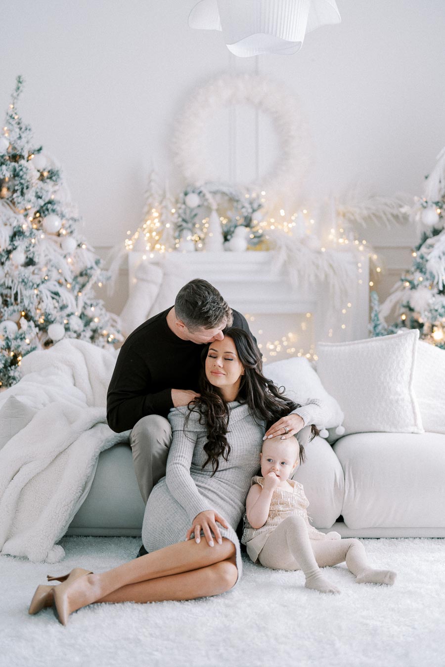 A loving family of three enjoys a cozy holiday moment on a white sofa, surrounded by festive Christmas decorations and twinkling lights.