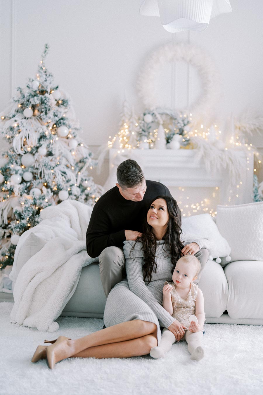 Family sitting on a white couch in a festive living room, with a decorated Christmas tree and twinkling lights, capturing a warm holiday moment.