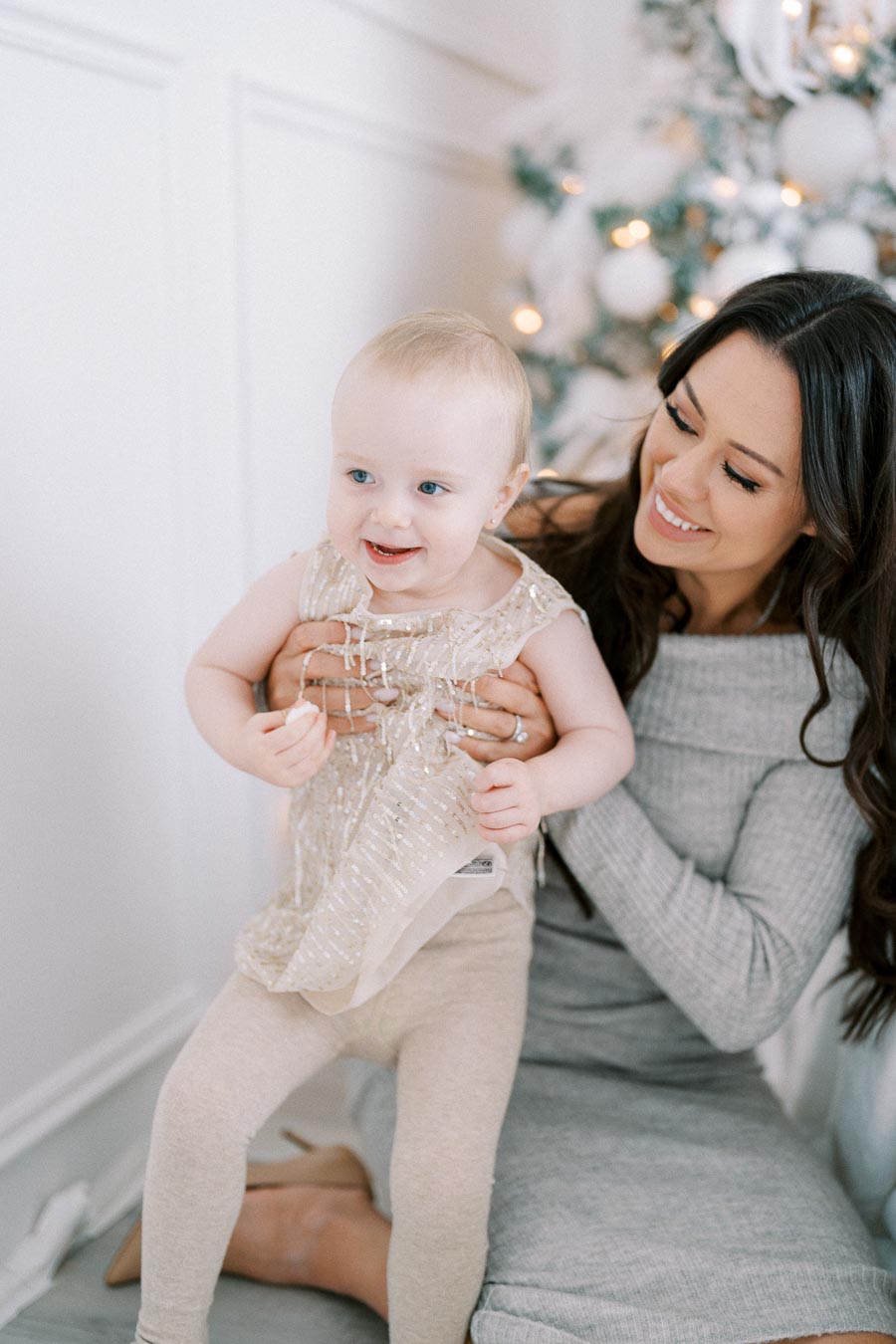 Mother holding her smiling baby dressed in gold near a decorated Christmas tree, creating a warm and joyful holiday moment.