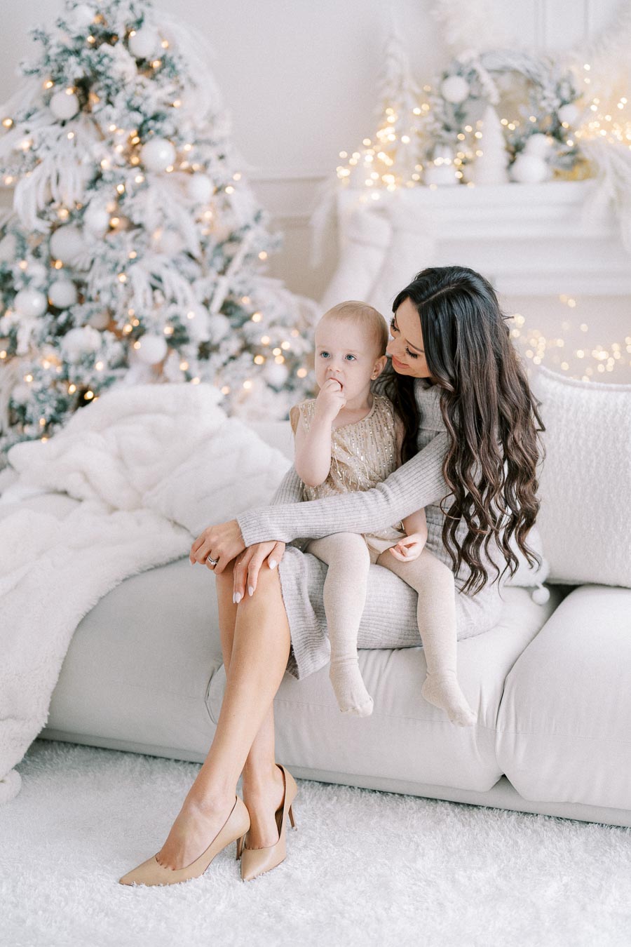 A mother and toddler sitting on a white sofa in a festive living room with Christmas decorations, including a lit Christmas tree and decorative garlands.