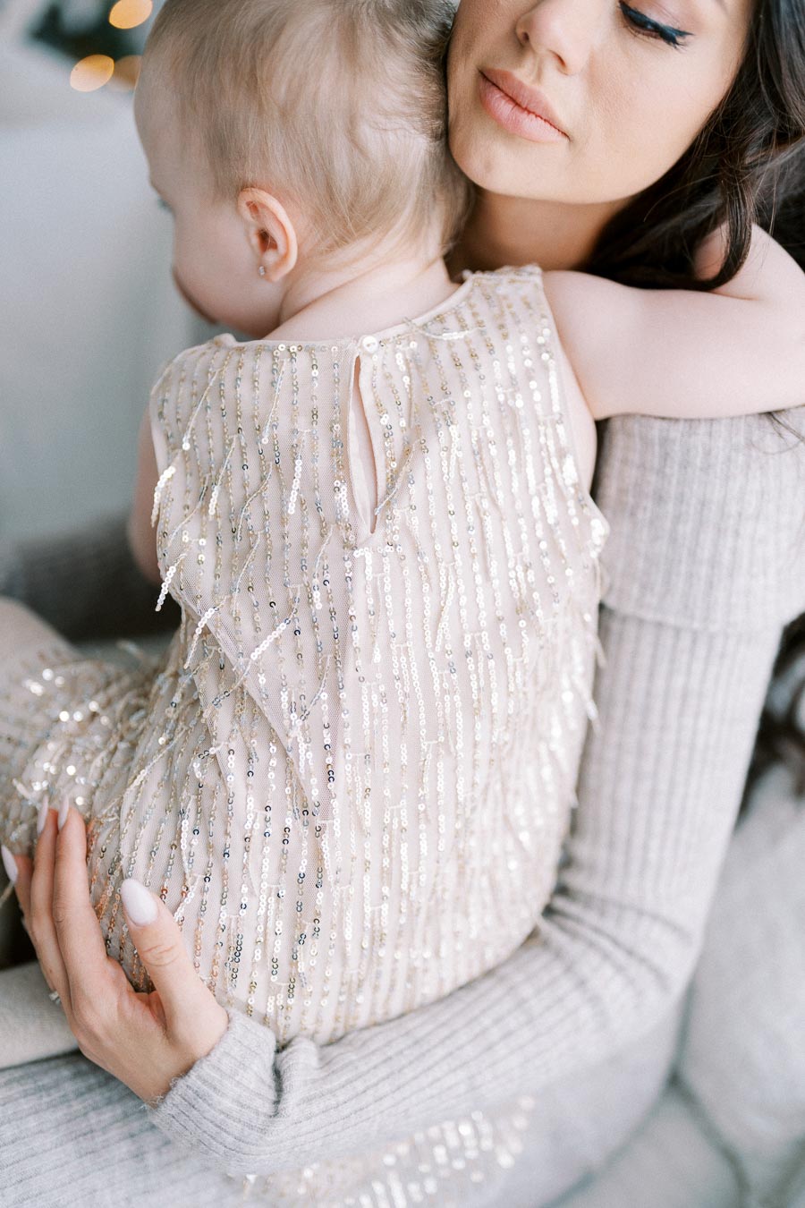 A mother holding her baby while sitting on a couch. The baby is wearing a sparkly dress with sequins, and the mother's expression appears gentle and serene. The cozy setting suggests a loving and warm atmosphere.