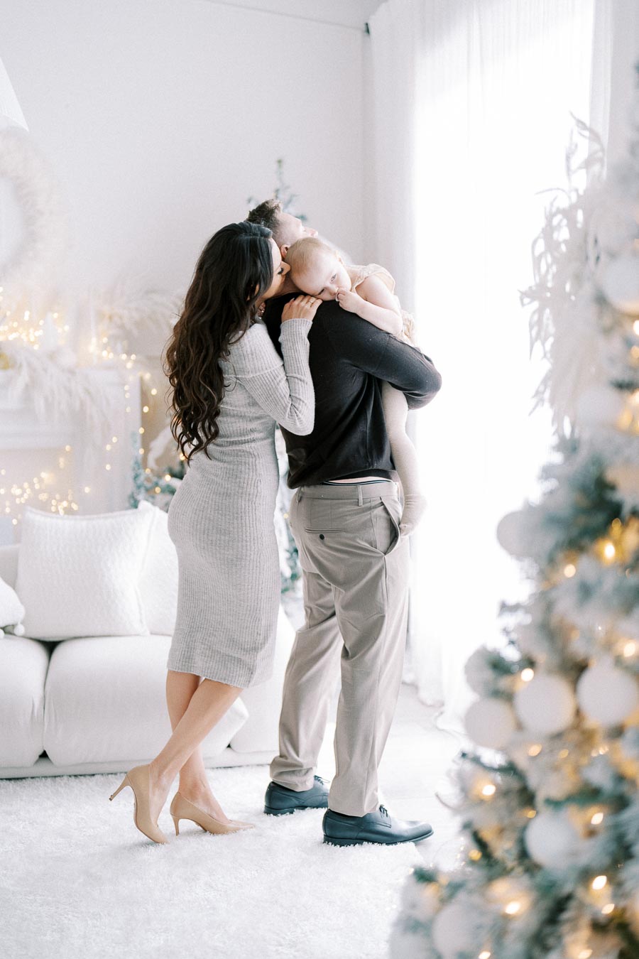 A family enjoying a cozy Christmas moment in a beautifully decorated living room, with a couple embracing and holding their child near a lit Christmas tree.