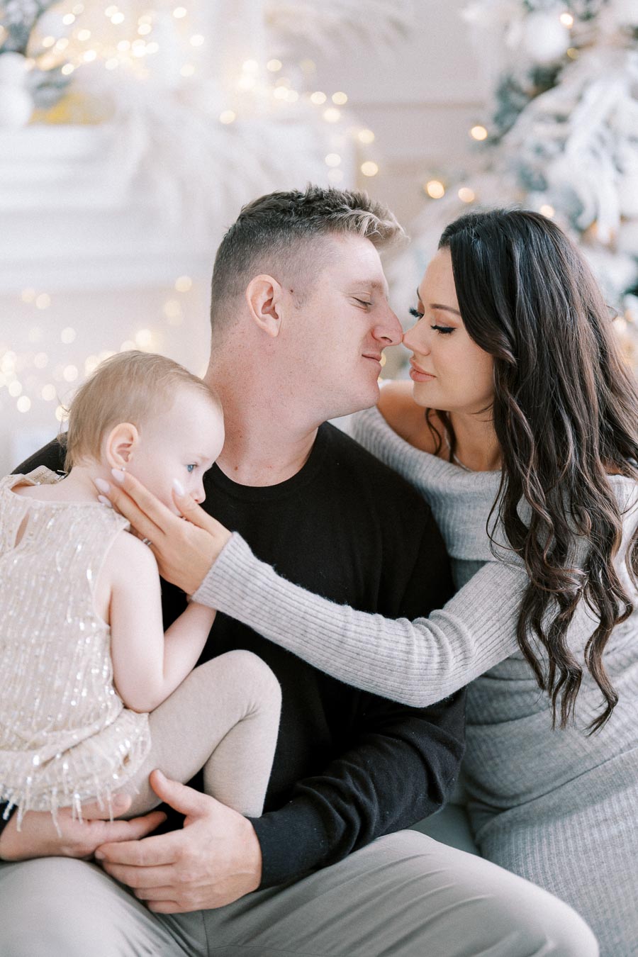 A loving family moment during the holiday season, with a couple embracing while holding their young child in front of a beautifully decorated Christmas tree, adorned with lights and ornaments.