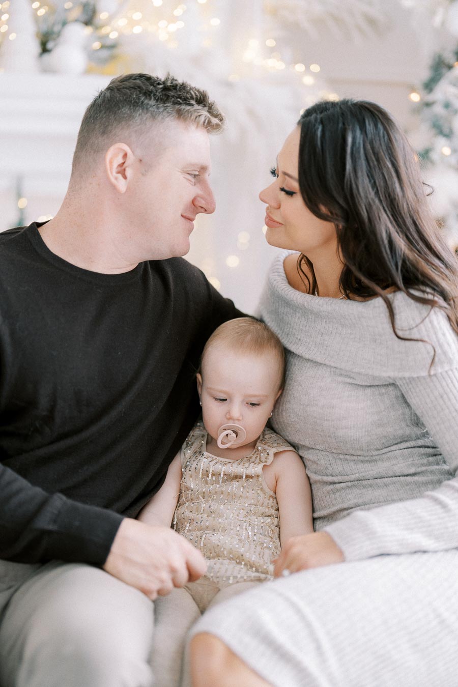 Family sitting together in a cozy setting, with warm lighting and festive decorations in the background. The parents look lovingly at each other while holding their baby, who is wearing a sparkly outfit with a pacifier.