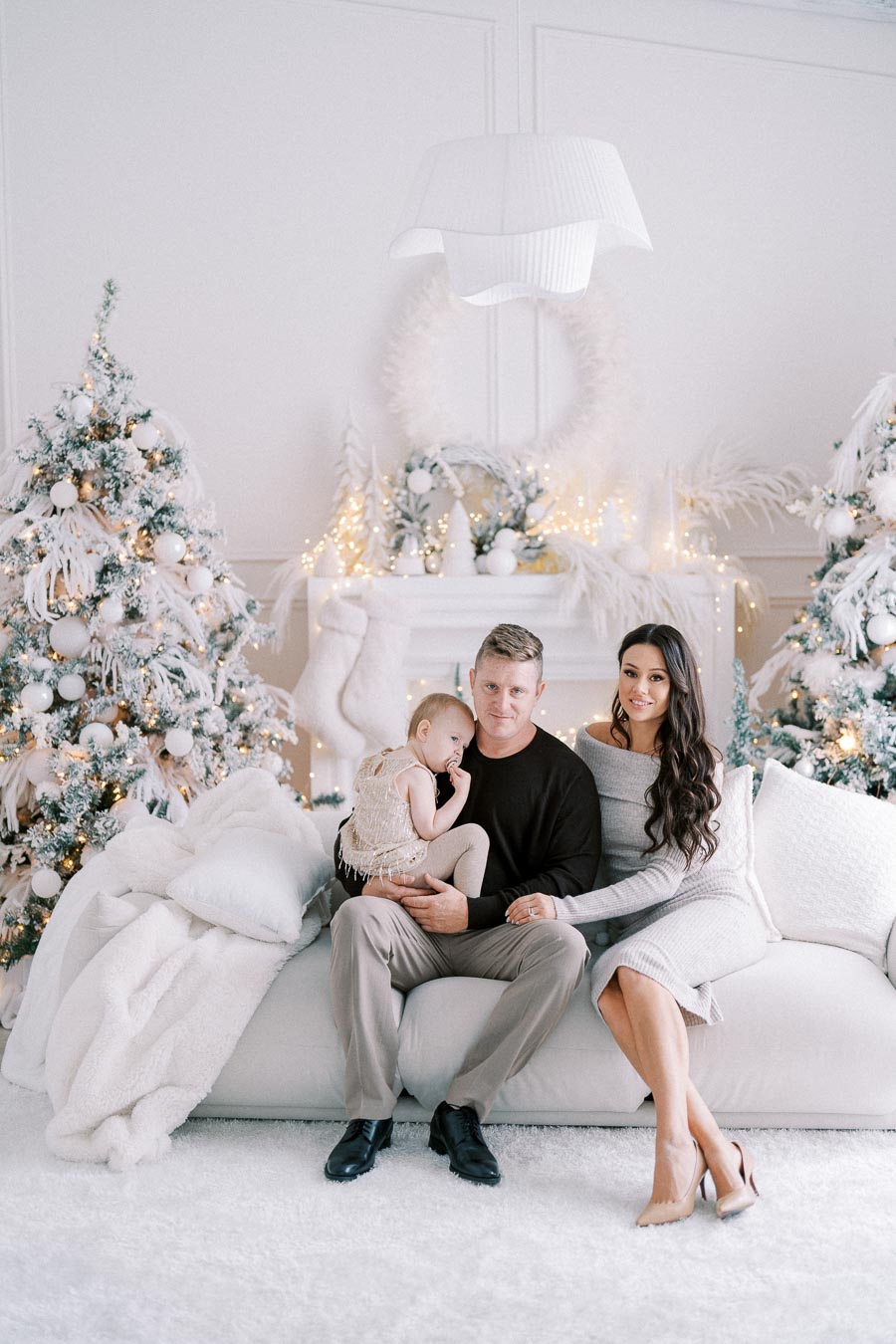 Family sitting on a white couch in a beautifully decorated living room with a winter theme; surrounded by snow-covered Christmas trees, twinkling fairy lights, and festive decor.