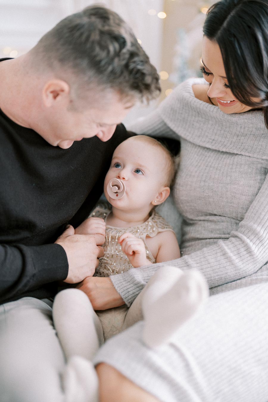 A loving family moment with parents smiling at their baby, who is lying on their lap with a pacifier, wearing a sparkly dress in a cozy home setting.