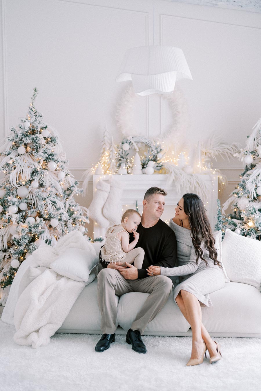 A family sitting on a plush white sofa, decorated for the holidays with snowy Christmas trees and warm lights in the background, creating a cozy winter atmosphere.