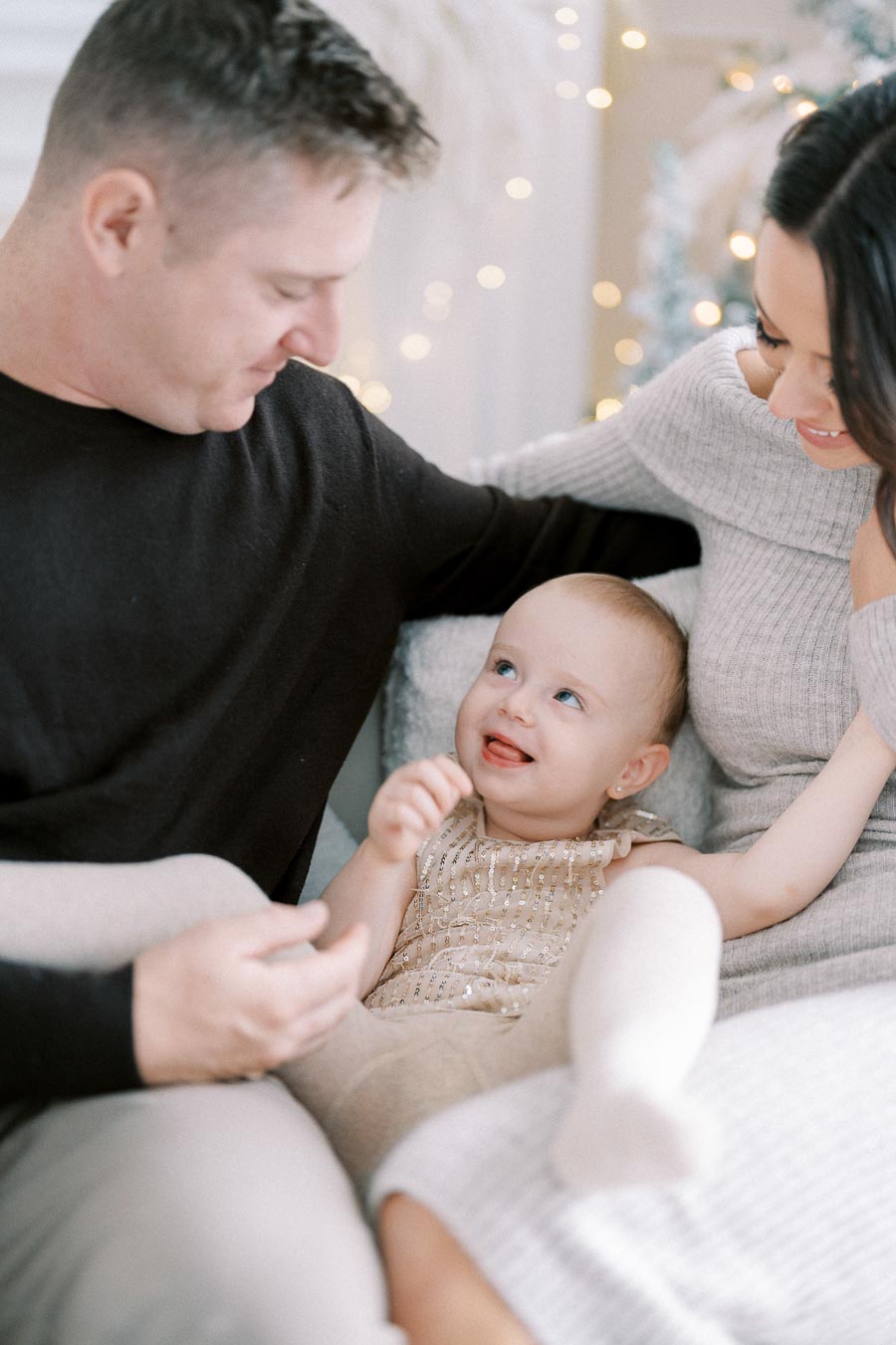 Happy family enjoying quality time together with a smiling baby wearing a sparkly outfit, surrounded by soft focus lights and cozy atmosphere.