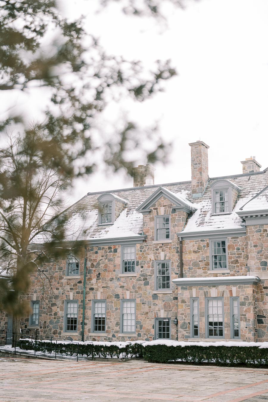 A large, classic stone house with snow-dusted roof and windows, surrounded by trees and hedges on a cold winter day.