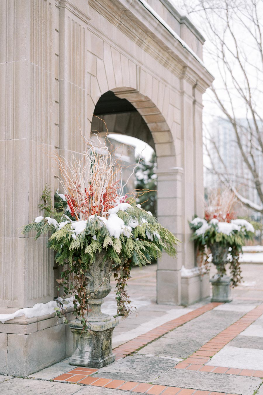 Stone archway decorated with snow-covered evergreen arrangements and red berries in ornate planters, capturing a wintery outdoor scene in a city setting.