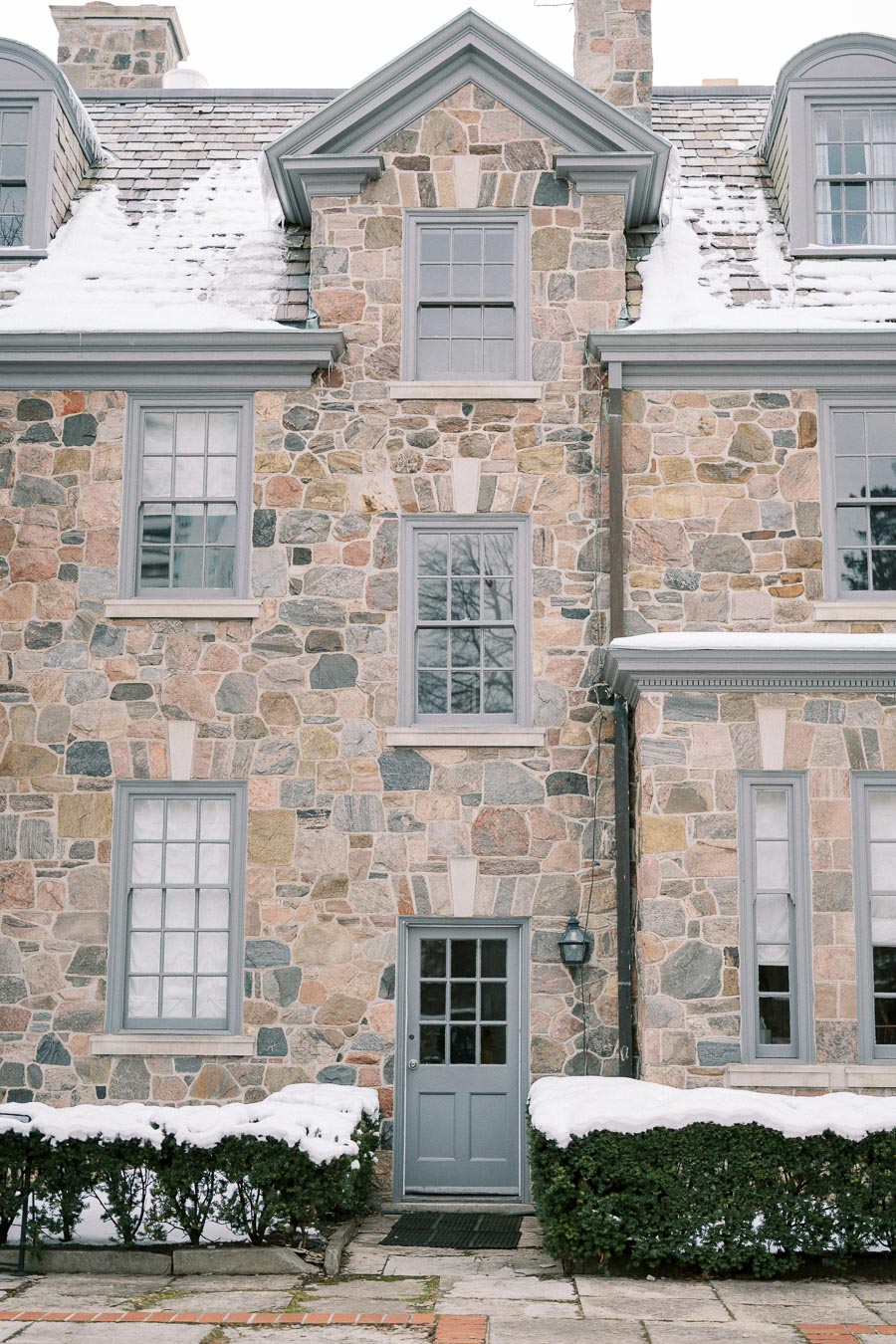 Front view of a charming stone house with gray window frames and a door, partially covered in snow, featuring a traditional architectural style.