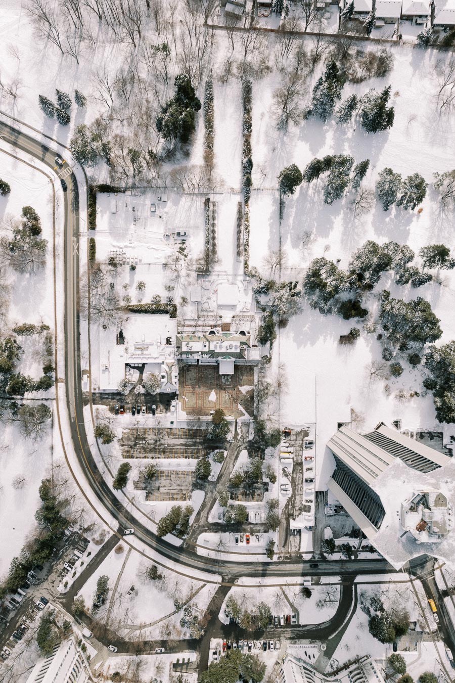 Aerial view of a snowy landscape featuring a winding road, trees, and buildings. The scene is covered in a blanket of white snow, creating a serene winter atmosphere. The layout of roads and structures appears carefully organized within the natural setting. Ideal representation of winter city planning and snowy urban architecture.