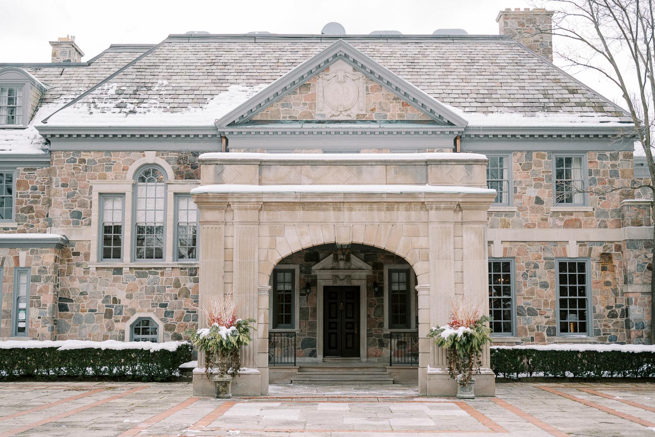 Elegant stone mansion with arched entryway, snow on the roof, winter landscape, and decorative planters on either side of the entrance.