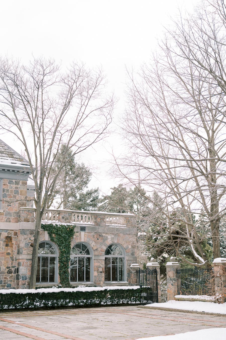 Elegant stone building with large arched windows surrounded by snow-covered trees and a wrought iron fence, capturing a serene winter scene.
