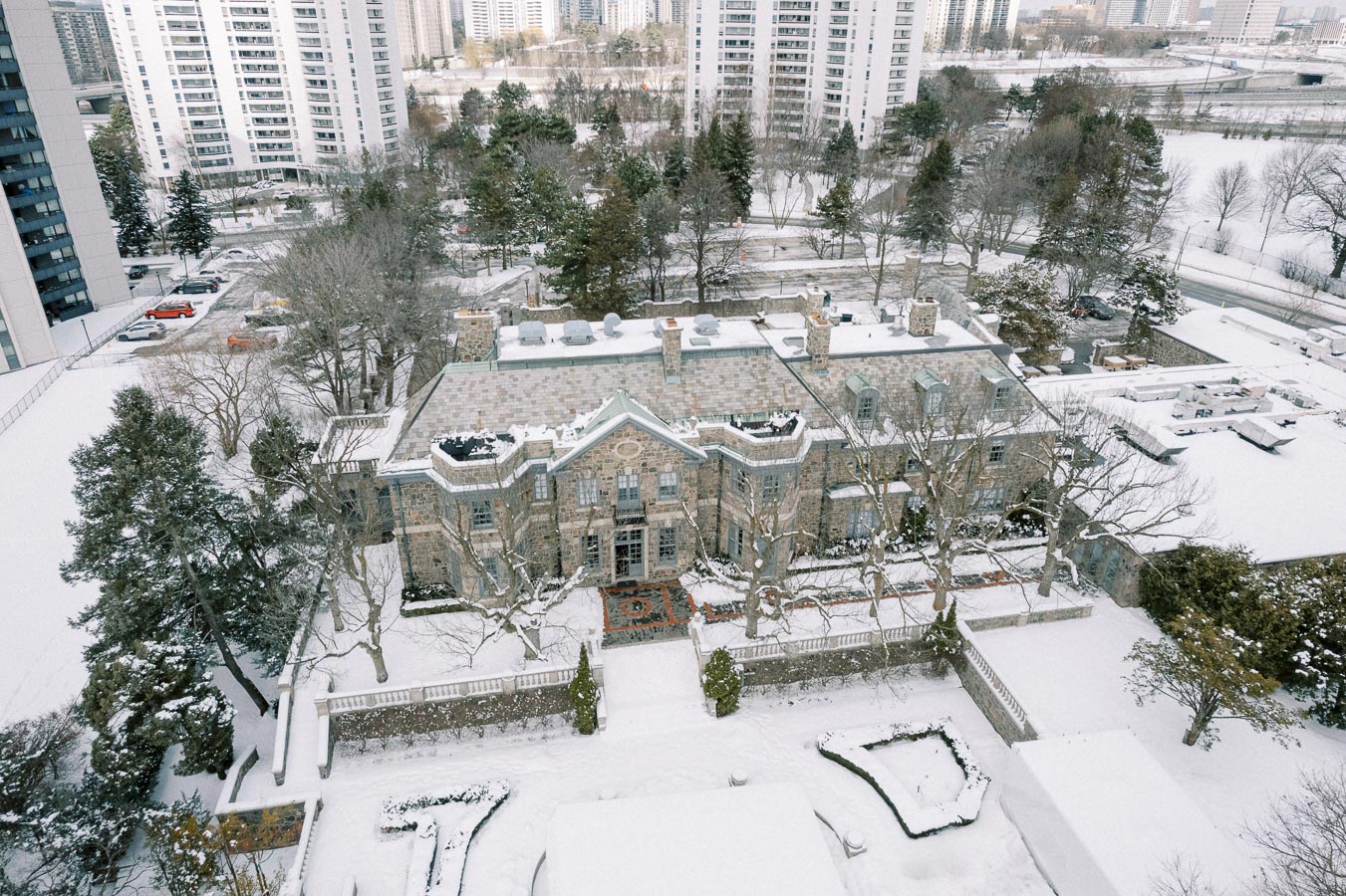 Aerial view of a snow-covered mansion surrounded by trees, set against a backdrop of urban high-rise buildings in the winter.