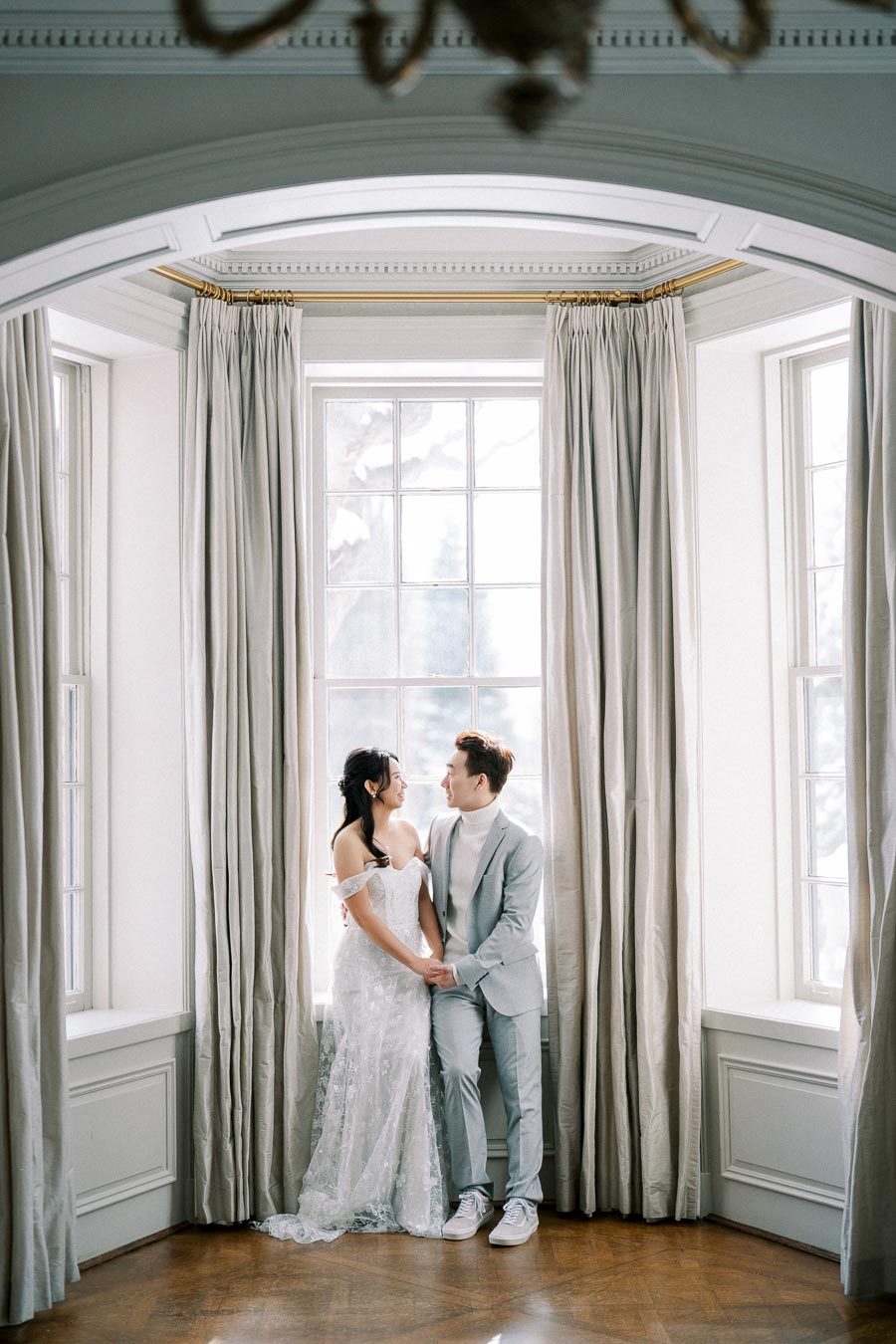 Wedding couple smiling and holding hands by a large window with elegant drapes, capturing a romantic moment in a bright and airy setting.