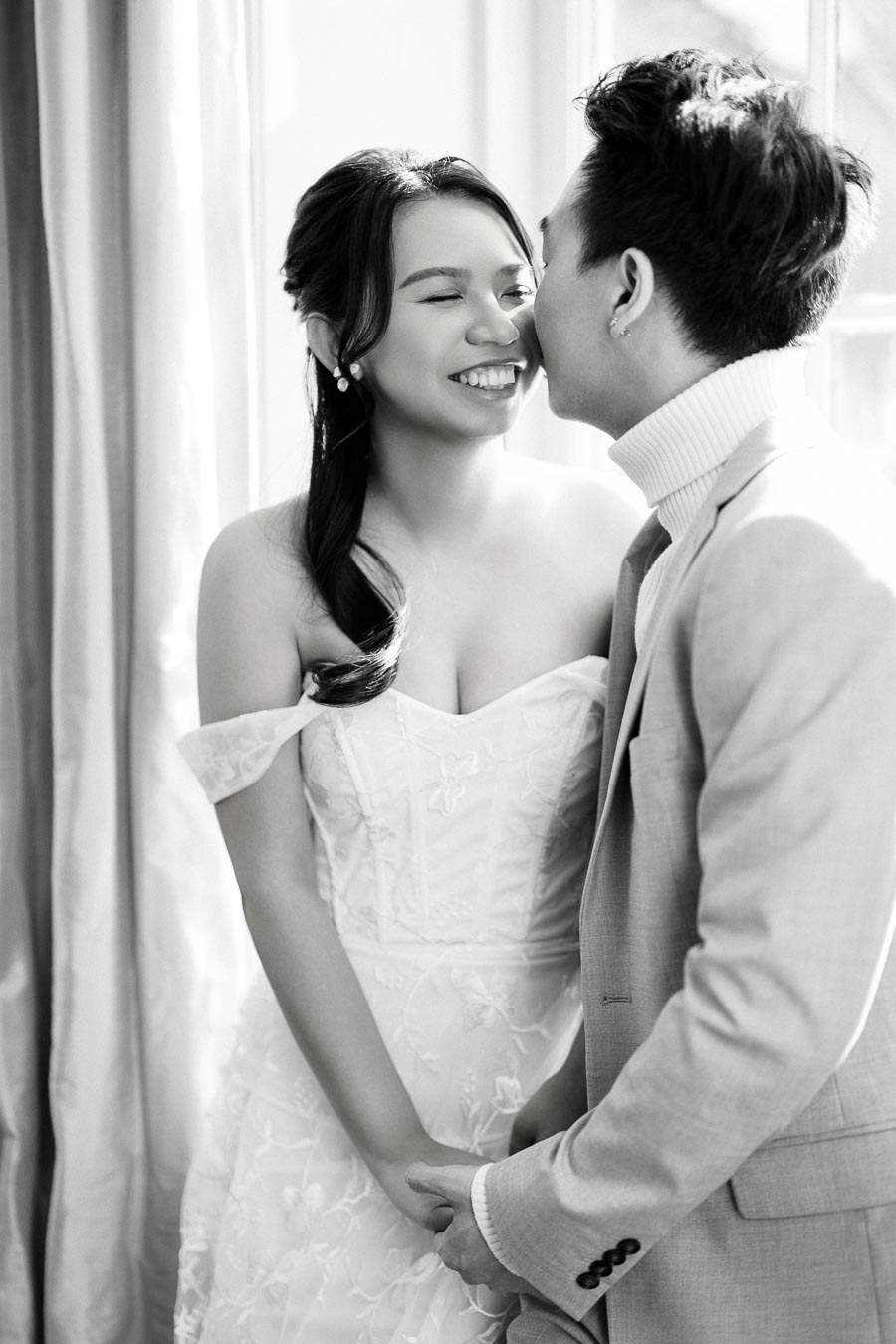 A couple sharing a joyful moment during their wedding, with the bride in an elegant white dress and the groom in a tailored suit, captured in a sunlit room.