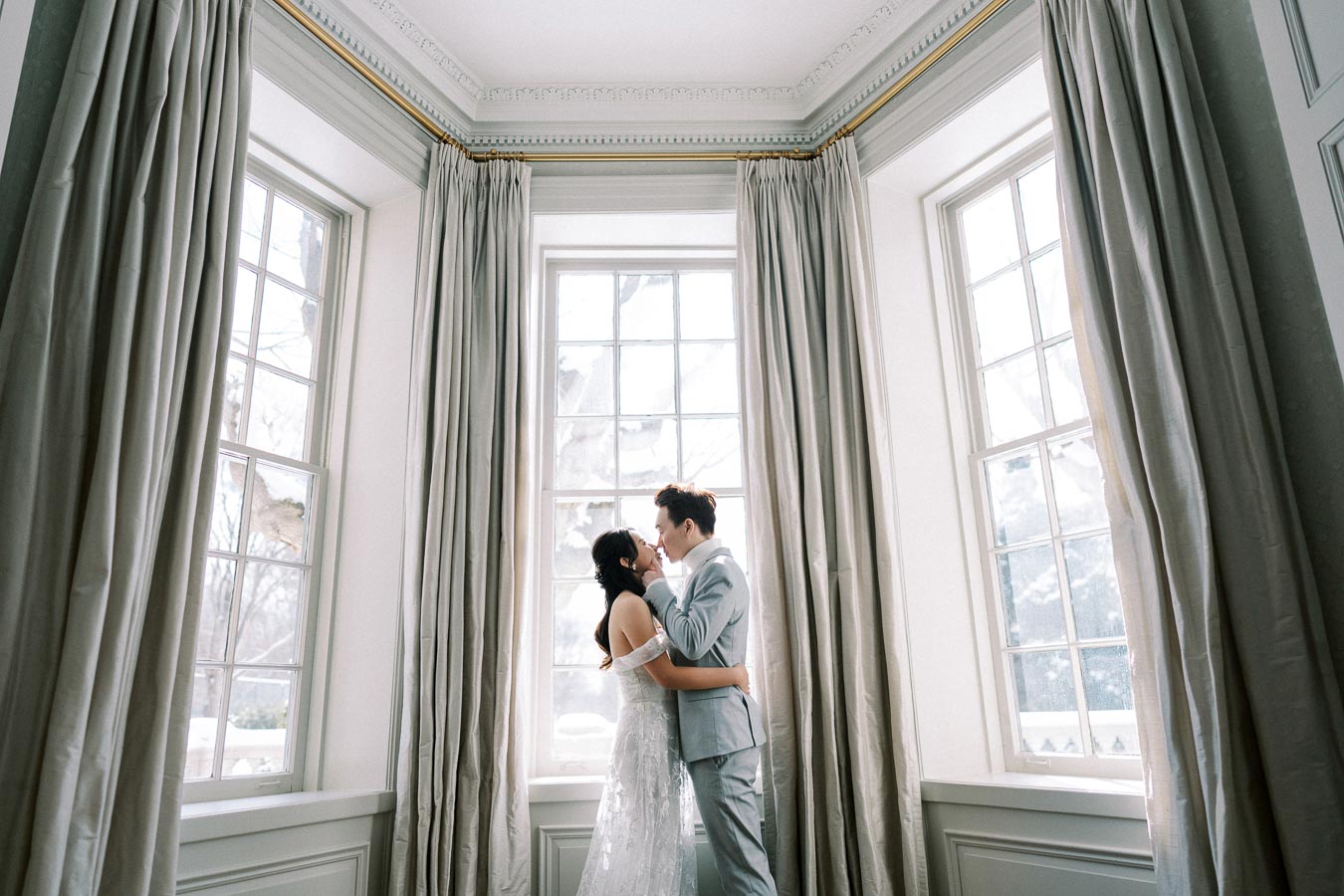 A romantic couple embraces in an elegant room with tall windows and flowing curtains, basking in natural light.