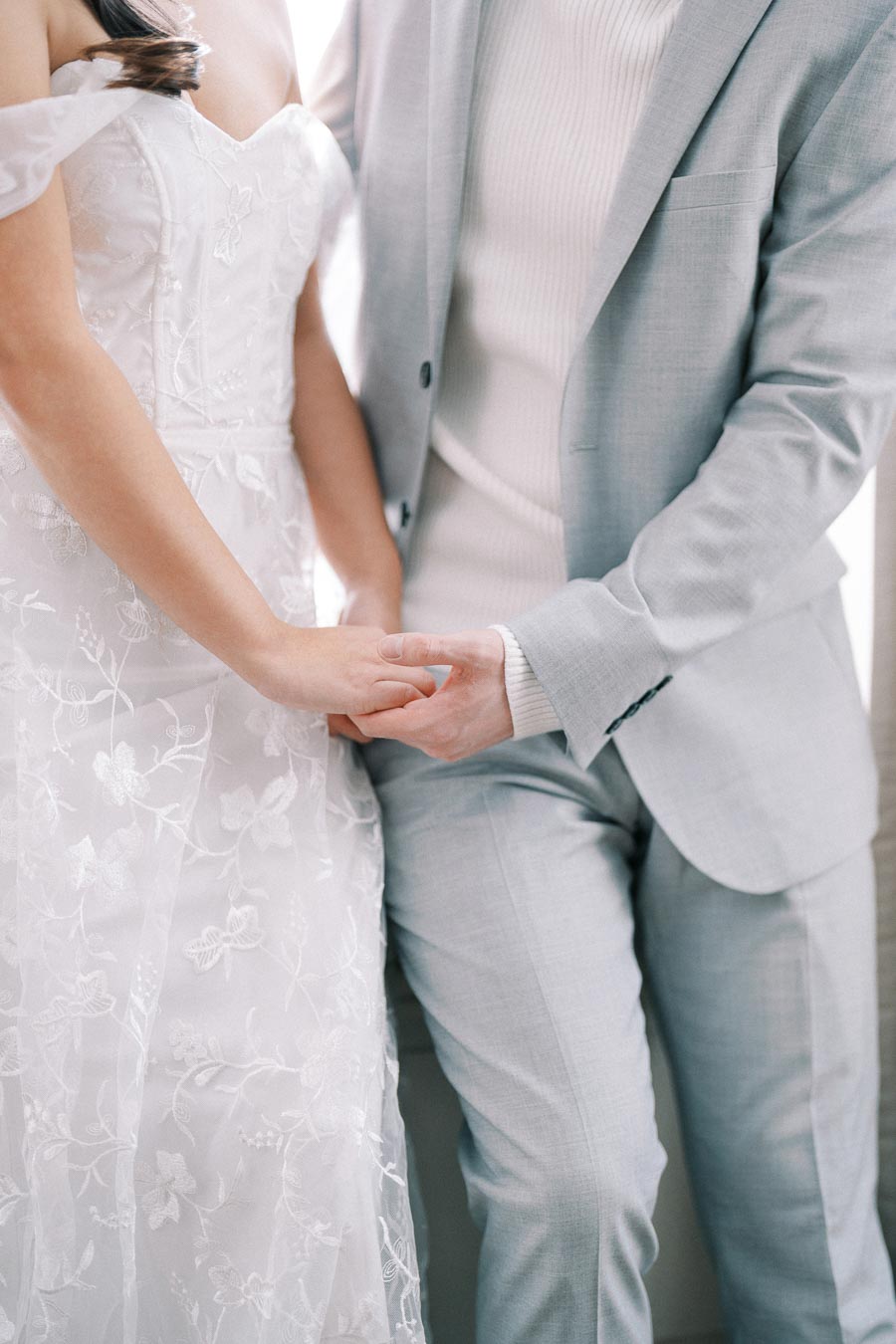 Bride and groom holding hands, bride in elegant floral wedding dress and groom in light grey suit, capturing a romantic and intimate moment.