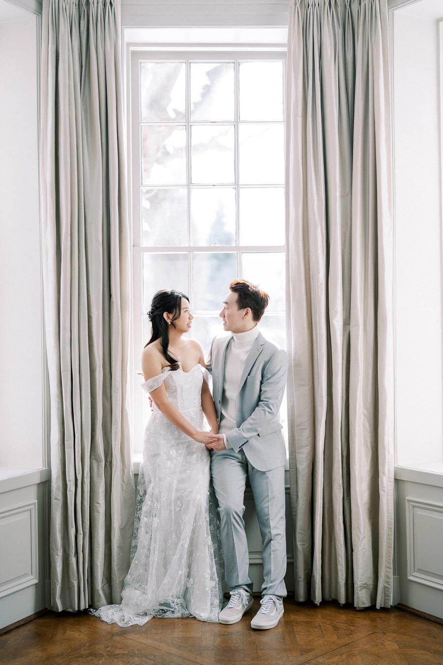 Elegant couple in wedding attire holding hands, standing by a large window with flowing curtains, natural light creating a romantic setting.