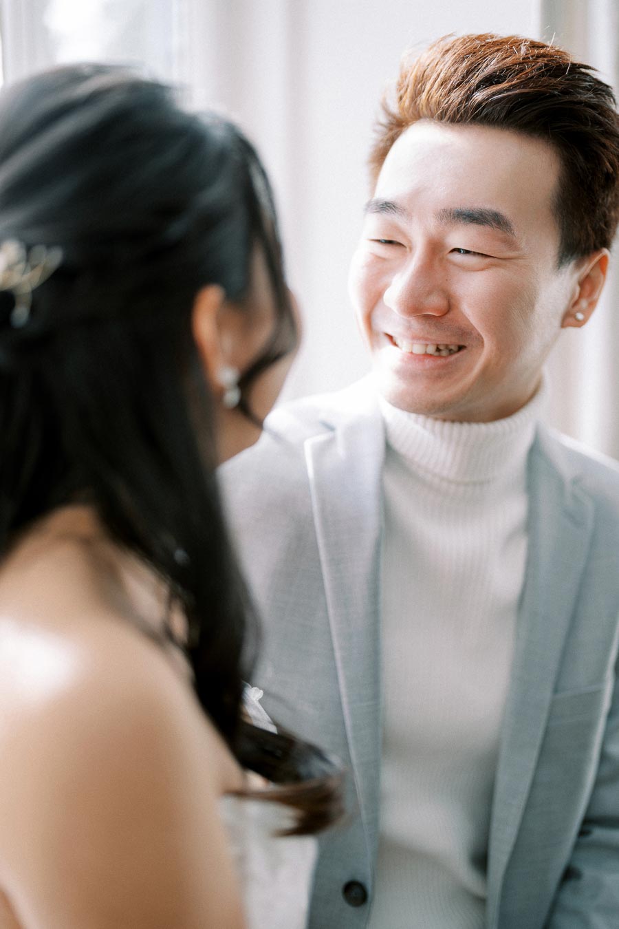 Smiling couple on wedding day, groom in light grey suit and bride with braided hair, sharing a joyful moment indoors.