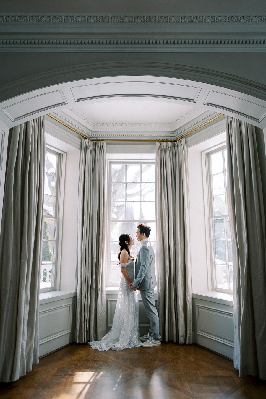 Elegant couple in wedding attire embracing by a large window in a beautifully decorated room with natural light flowing through curtains