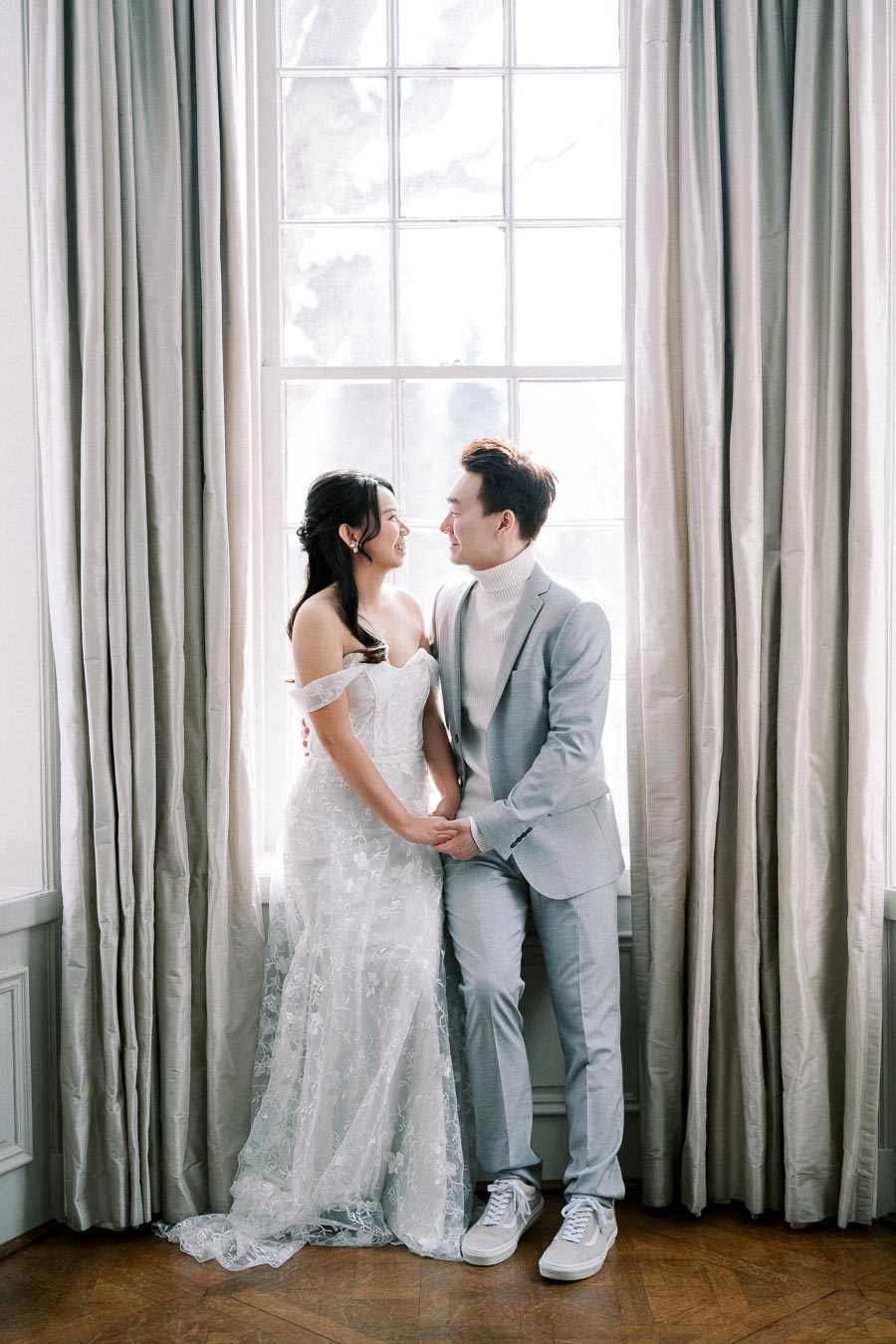 Elegant couple in formal attire standing by a large window with soft curtains, embracing and smiling at each other, captured in a bright indoor wedding setting.