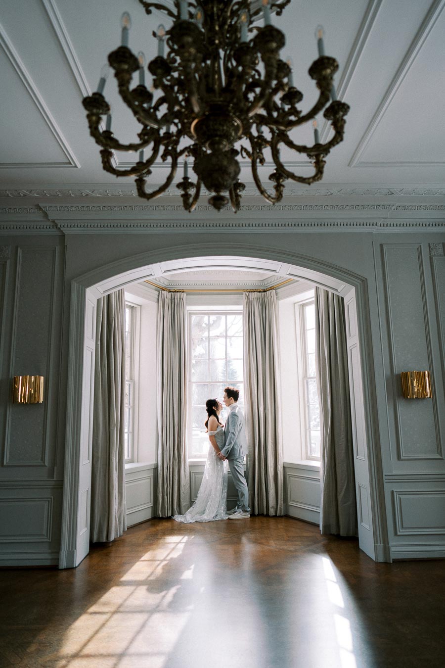 Elegant couple in wedding attire embracing by a large window in a beautifully decorated room with soft natural lighting and a classic chandelier.