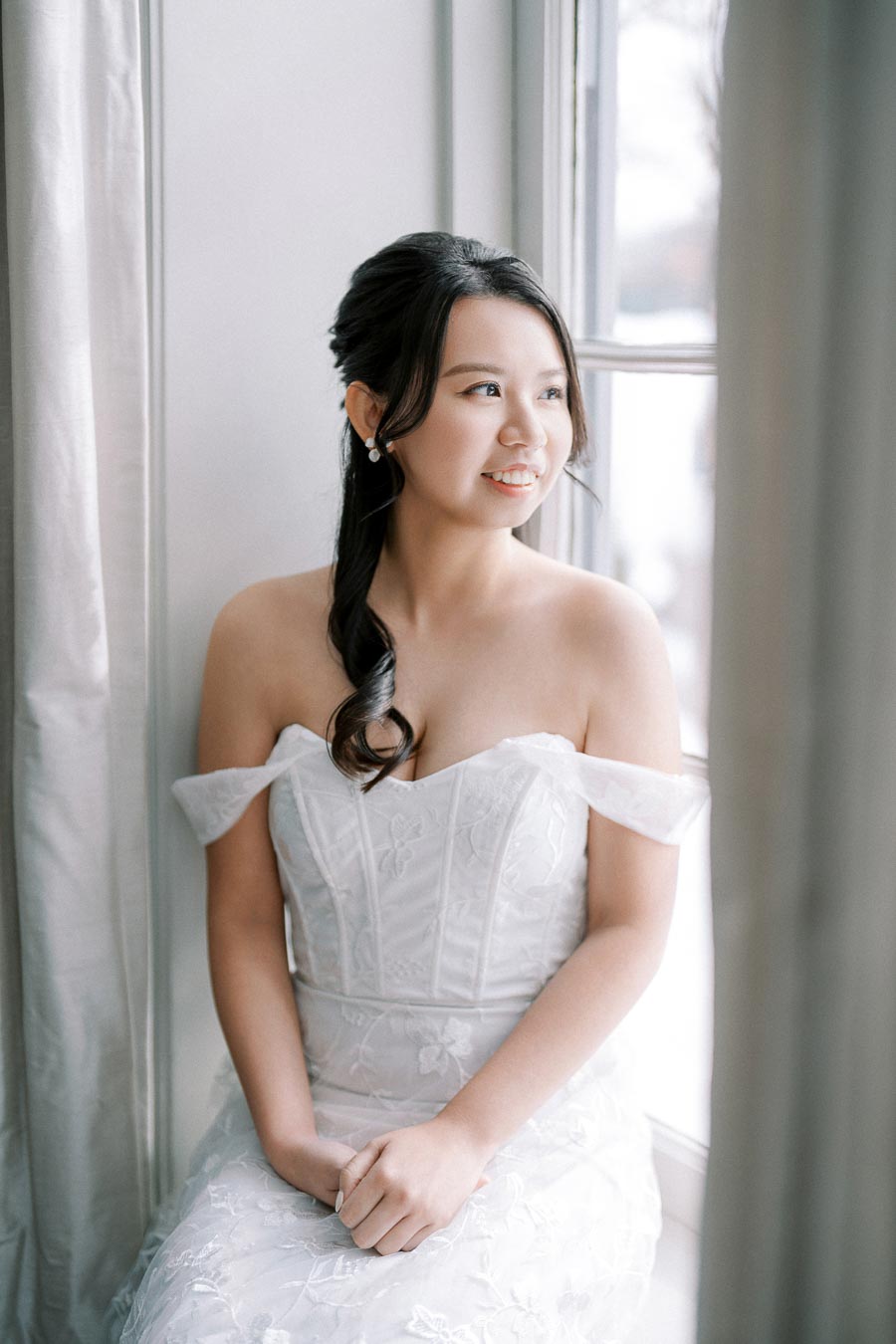 Bride in elegant white wedding dress sitting by a window, looking outside with a serene expression, natural lighting highlighting the lace details of the dress.