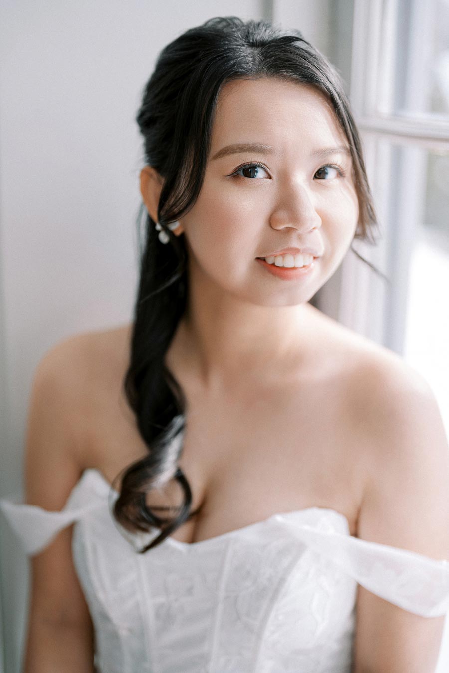 Elegant bride smiling in natural light, wearing a white off-shoulder wedding dress with intricate lace details and long dark hair styled in a braid. Perfect bridal portrait showcasing timeless beauty and grace.