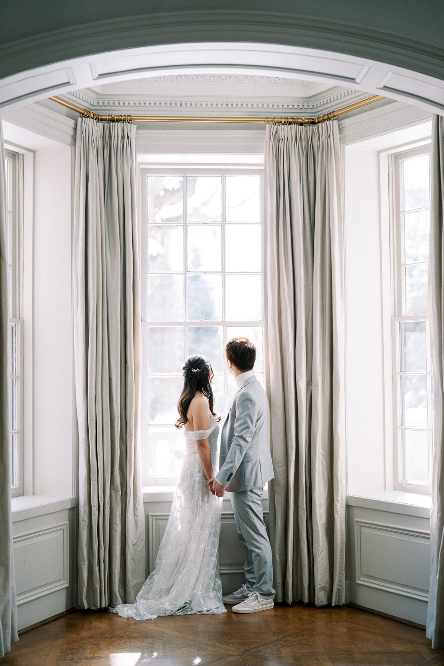 A bride and groom stand hand in hand, gazing out a large window framed by elegant curtains, showcasing a serene and romantic setting on their wedding day.