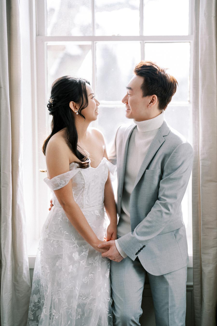 A happy couple standing together in front of a large bright window. The woman is wearing a delicate white lace dress, and the man is dressed in a light gray suit with a turtleneck sweater. They are holding hands and gazing at each other lovingly, creating a romantic atmosphere.