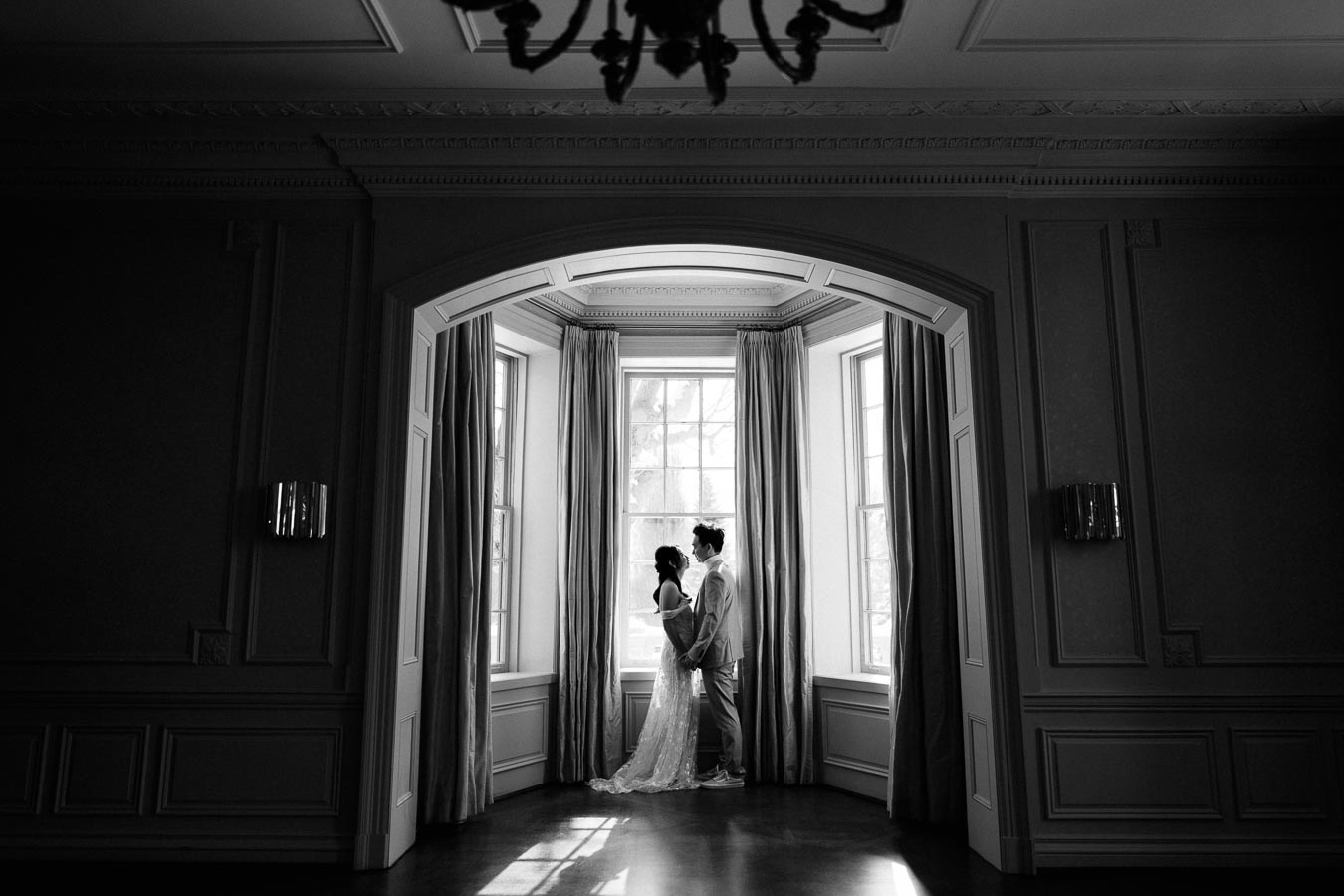 Black and white image of a couple standing in a sunlit window alcove, embracing and gazing at each other, creating a romantic and elegant ambiance in a classic room with high ceilings and intricate molding.