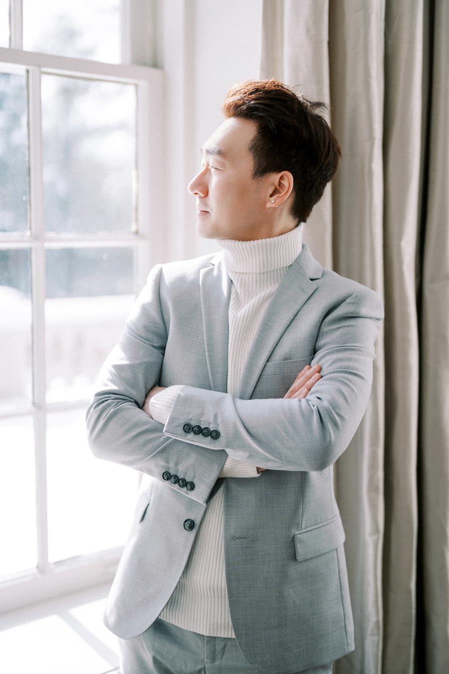A man in a light gray suit and white turtleneck sweater stands with arms crossed, gazing out the window. The room is softly lit with natural daylight, highlighting a serene and contemplative atmosphere.