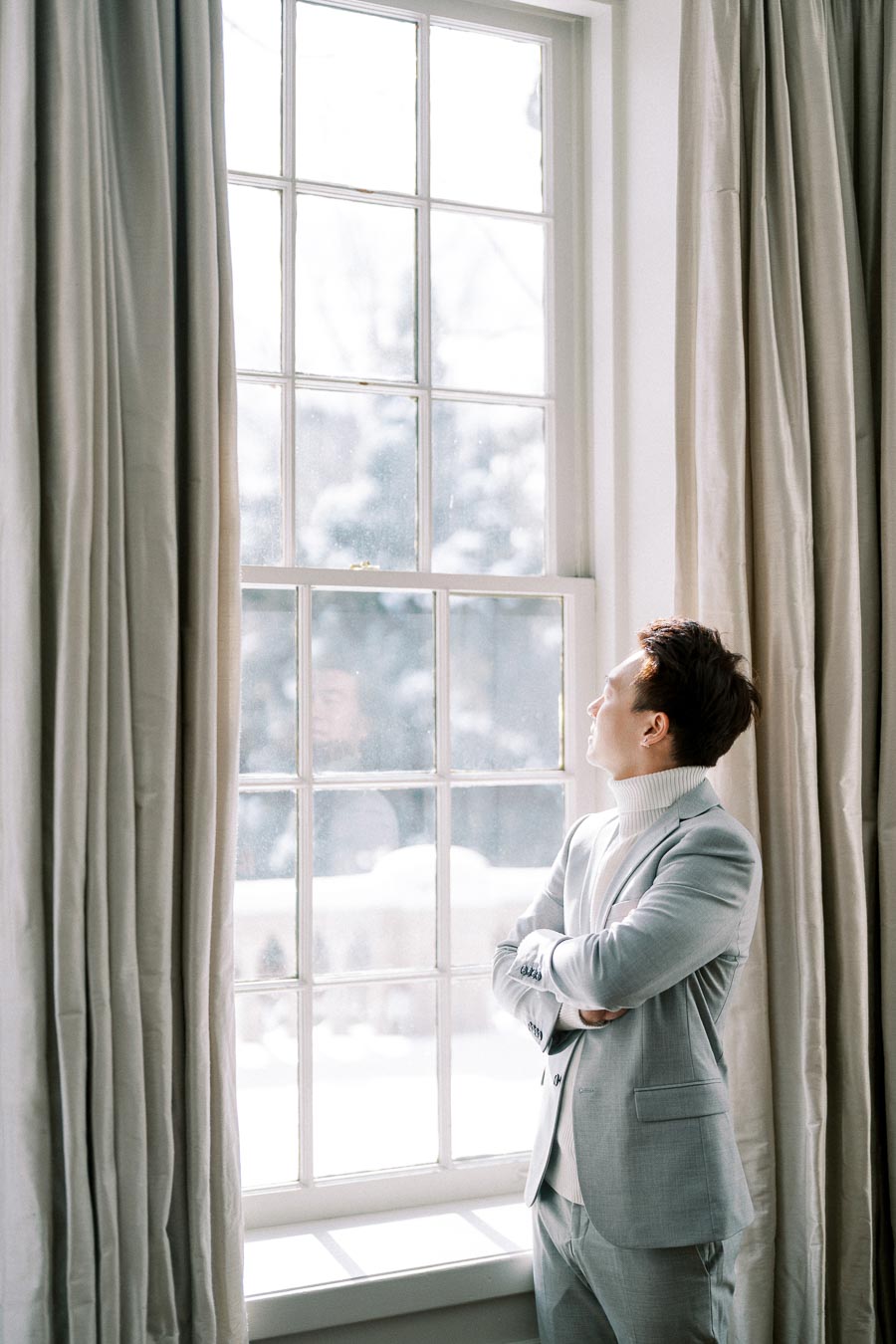 A man in a light gray suit stands with arms crossed, gazing thoughtfully out a large window in a softly lit room. Drapes frame the window, and the snowy landscape outside creates a serene atmosphere.