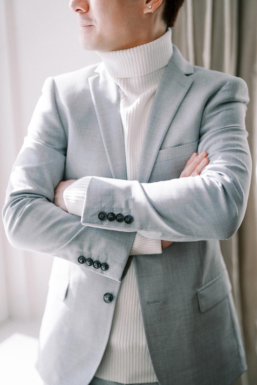 Man wearing a light gray suit and a white turtleneck sweater, standing with arms crossed, facing sideways.