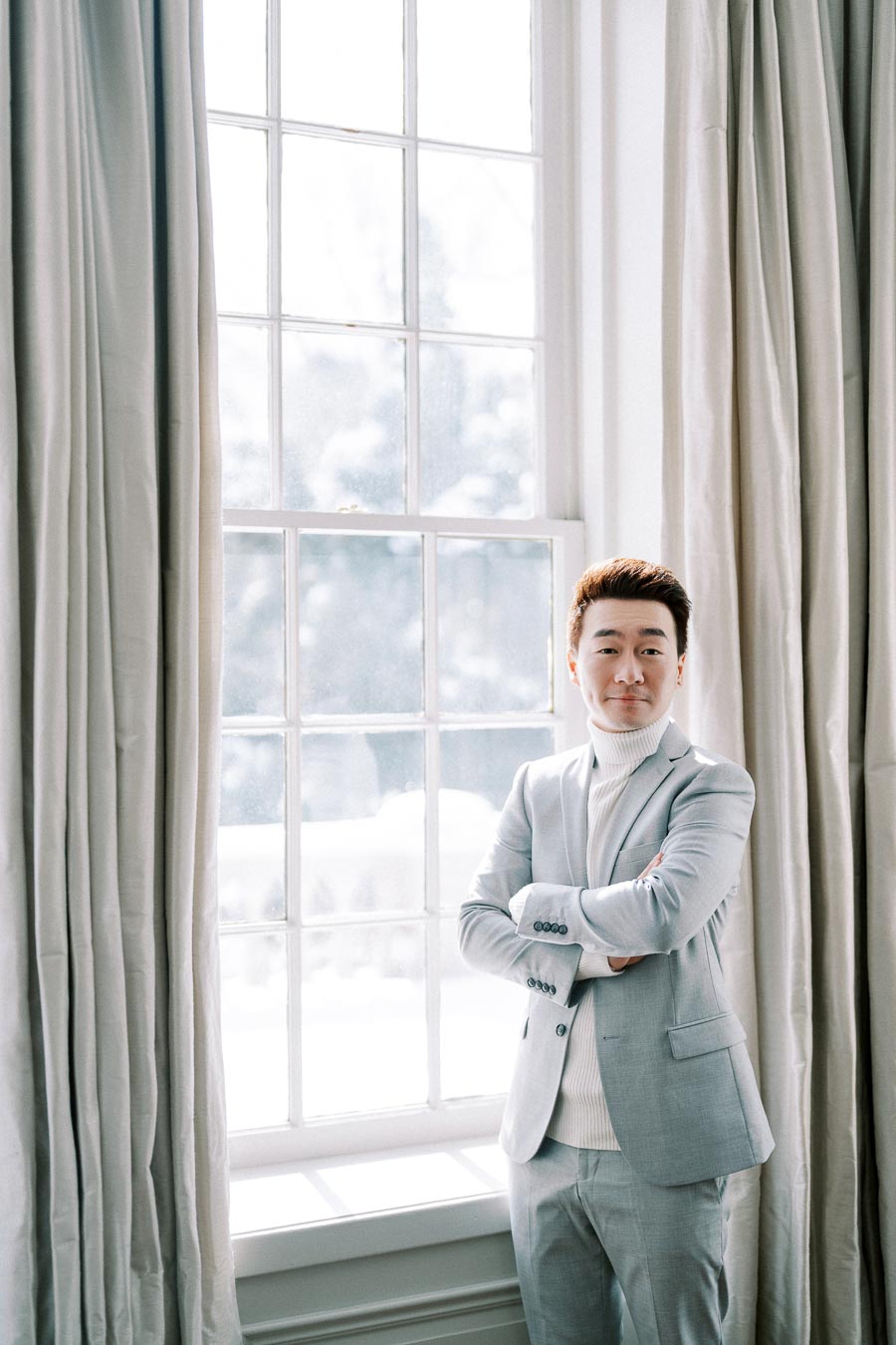 Man in light gray suit standing by a bright window, arms crossed, with curtains framing the scene.