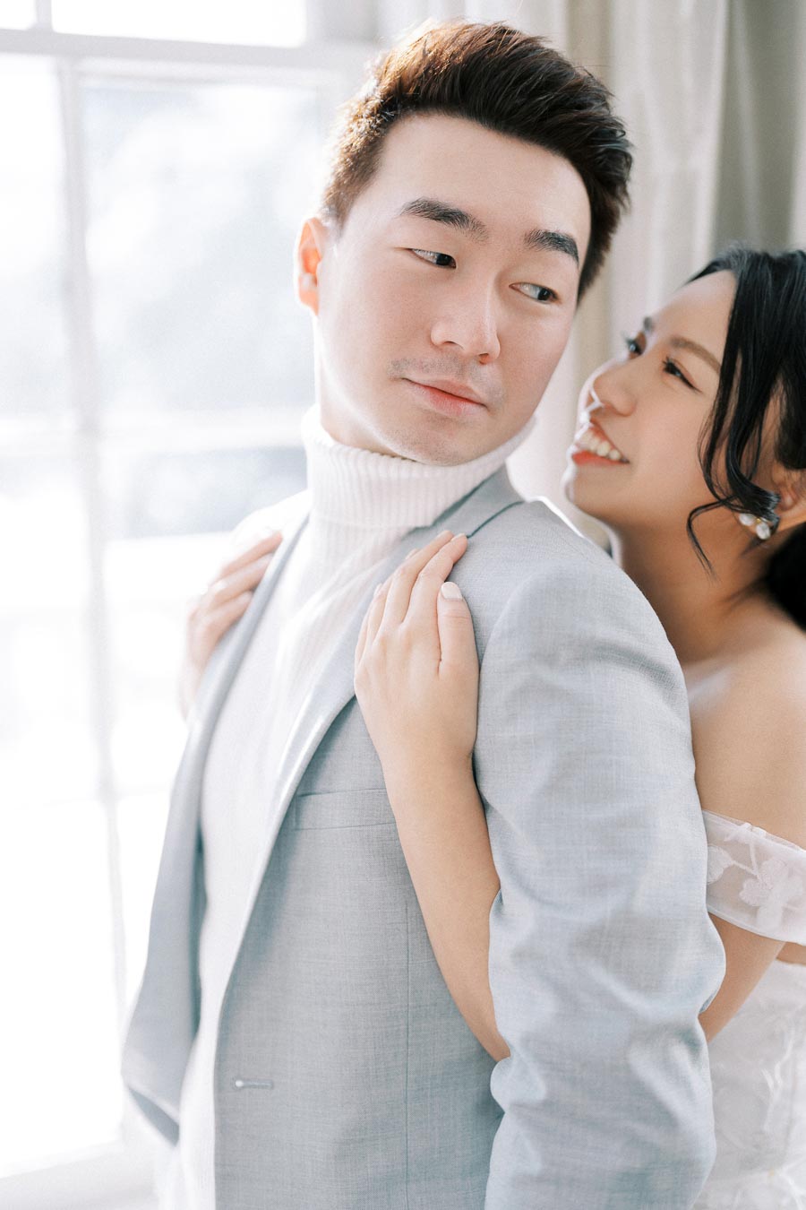 A romantic couple embracing by a window, with the woman smiling warmly as she leans on the man's back. The man is wearing a light gray suit with a white turtleneck, and the woman is in an off-shoulder white dress. The soft natural light creates an intimate and elegant atmosphere.