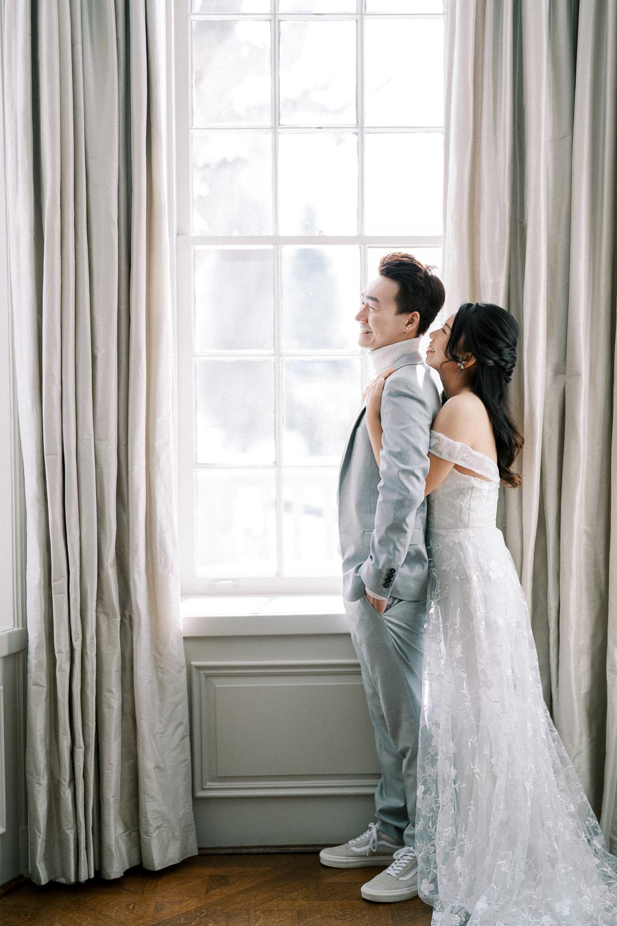 Wedding couple sharing a joyful moment by a sunny window, groom in a light gray suit and bride in an elegant off-shoulder lace gown, creating a romantic and serene atmosphere.