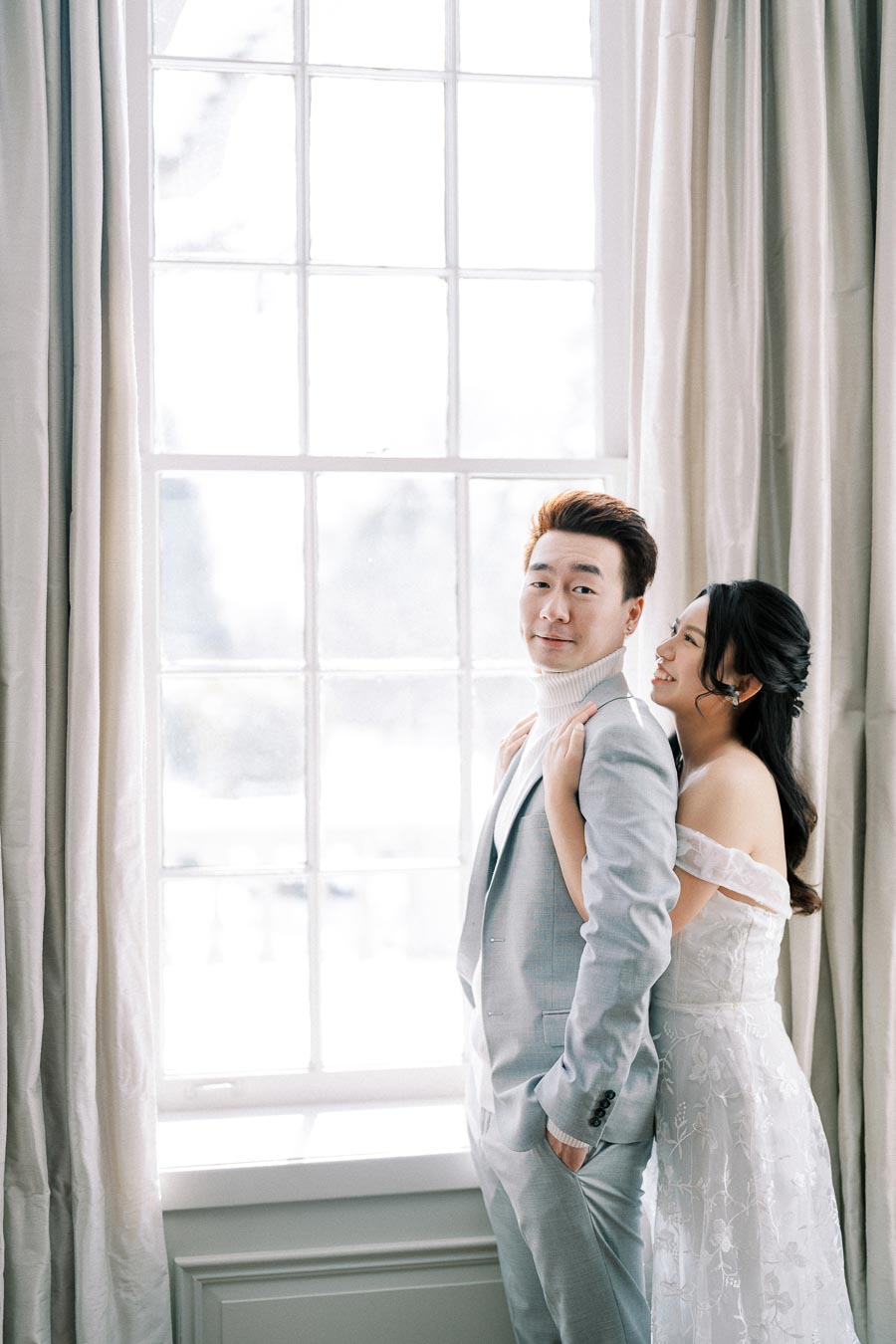 A happy couple poses by a large window in elegant wedding attire, with the groom in a light grey suit and the bride in an off-shoulder lace gown, creating a romantic atmosphere.